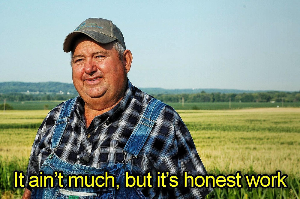 Meme photo of an older farmer in overalls and a cap standing in front of farm fields, captioned 'It ain't much, but it's honest work.'